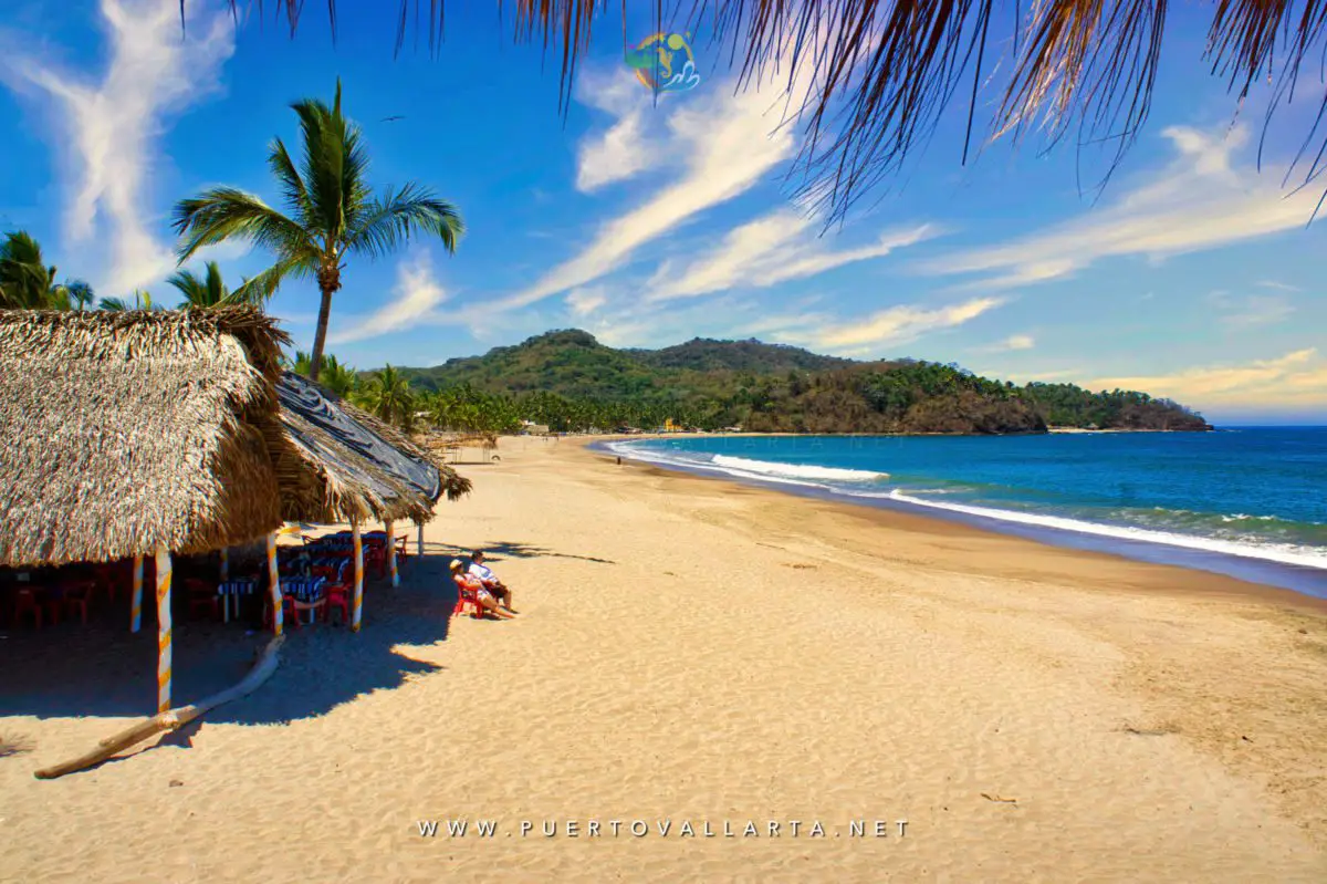 Thatched beach palapa with tables and chairs beside the curving shore at Lo de Marcos